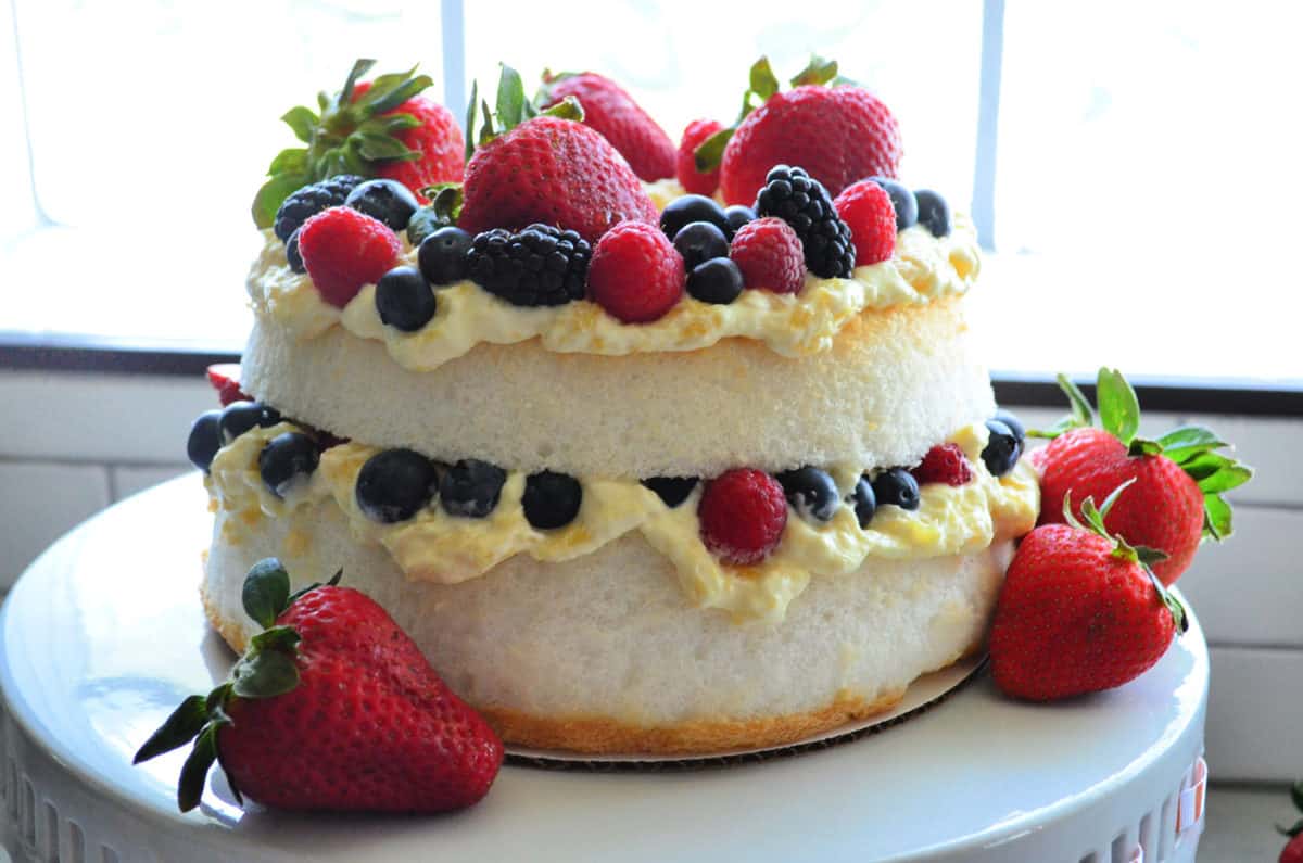 Two layer cake with yellow cream and berries on a white cake stand. 