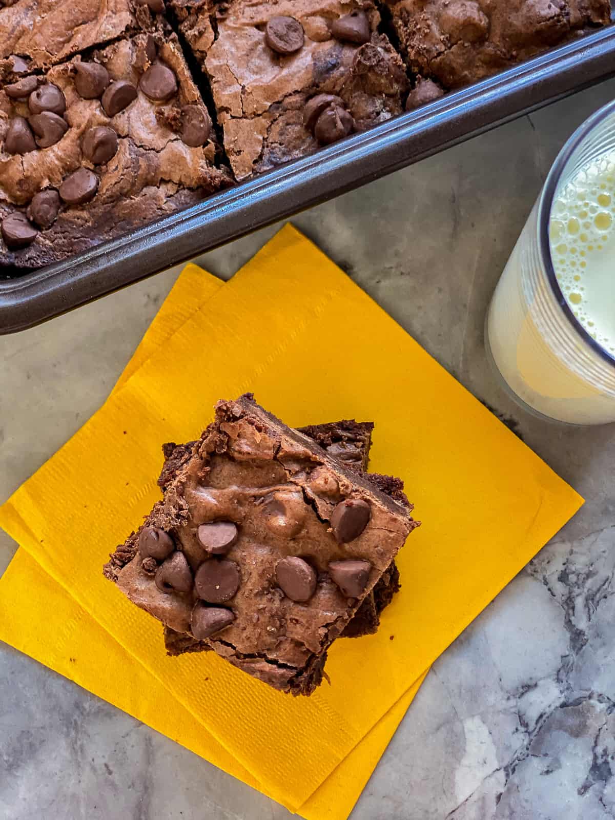 A yellow napkin with two square brownies on top, next to a glass of milk and a glass pan of brownies.