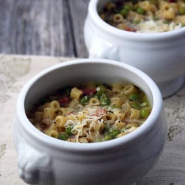 Two white urns full of peas, pasta, and meat in broth sittind on a wood board.