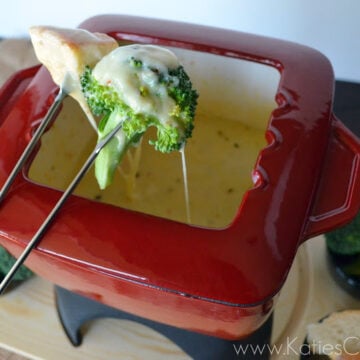 Fondu fork with broccoli dripping with cheese over red fondu pot filled with speckled melted cheese.