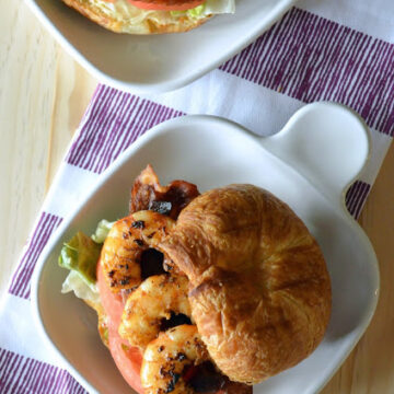 plated croissant sandwhich with top slightly moved to the side, revealing shrimp, lettuce, bacon, and tomato.