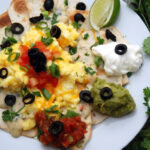 plated nachos topped with eggs, cheese, red salsa, guac, sour cream, cilantro, green onion, olive.