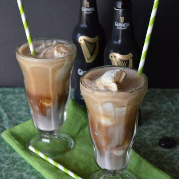 two sunday glasses containing brown creamy liquid with chunks of ice cream and paper straws with guinness bottles in background..