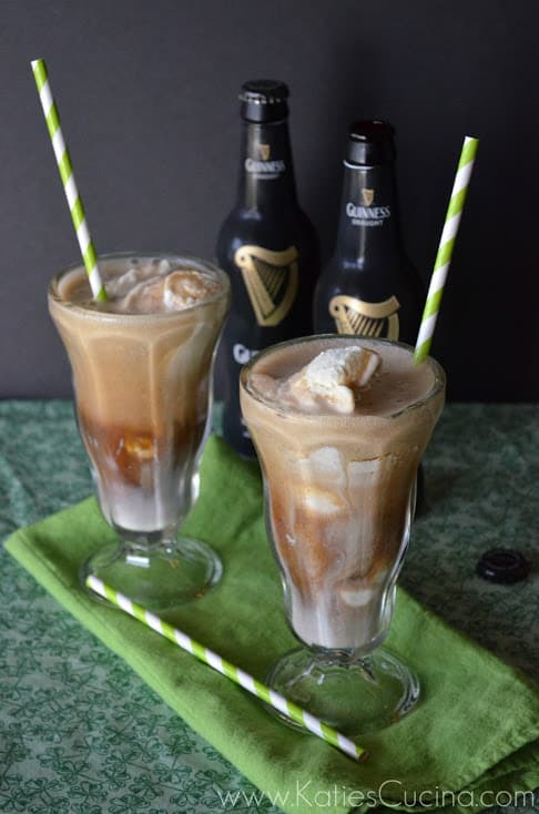 two sunday glasses containing brown creamy liquid with chunks of ice cream and paper straws with guinness bottles in background..