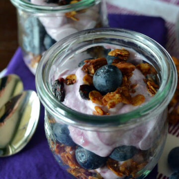 Two glasses filled with yogurt, granola, and fresh blueberries on a purple tablecloth.
