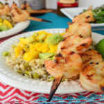 Grilled shrimp skewers plated over bed of roasted mango chipotle rice with lime slice on tablecloth.