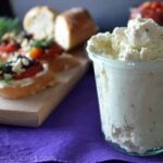Glass containing Garlic and Herb Whipped Feta with board of Greek Crostinis in background.