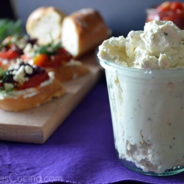 Glass containing Garlic and Herb Whipped Feta with board of Greek Crostinis in background.