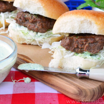 Close up of beef sliders on board with bowl of sauce and knife dripping with sauce.