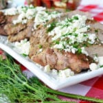 Plated grilled pork tenderloin topped with feta cheese and dill on picnic tablecloth.