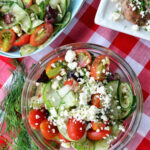 Top view greek cucumber, tomato, and feta salad artistically placed near dill on tablecloth.