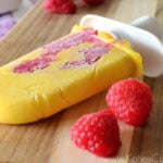 creamy orange colored popsicle with raspberries visibly frozen in it on board next to fresh raspberries.