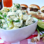 Bowl of Green Onion & Pea Potato Salad next to green onions and limes with sliders in background.