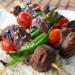 skewers of snow peas, beef, mushrooms, cherry tomatoes garnished with sesame seeds over white rice bed.