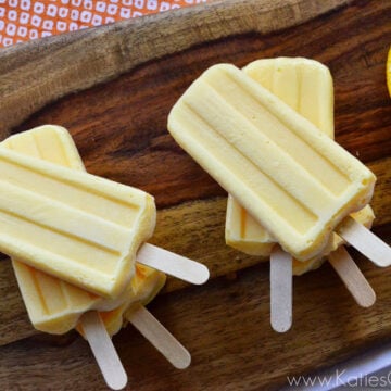 Two stacks of homemade light orange popsicles next to orange slices on wooden board.