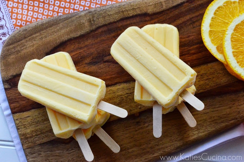 Two stacks of homemade light orange popsicles next to orange slices on wooden board.