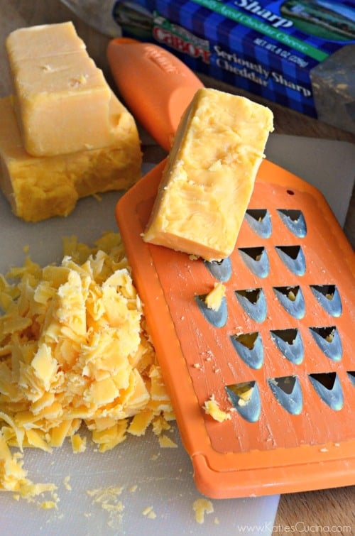 Orange grater with blocks of cheddar on the top and on the side with shredded cheddar cheese on cutting board.