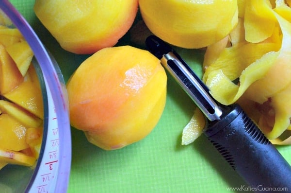 Peeled whole peaches on a green cutting board with a black peeler facing up. 