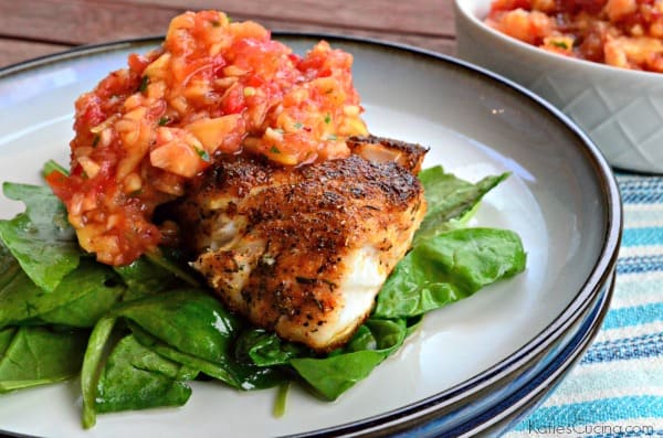 Blackened white fish topped with peach salsa resting on a bed of spinach on a white plate. 