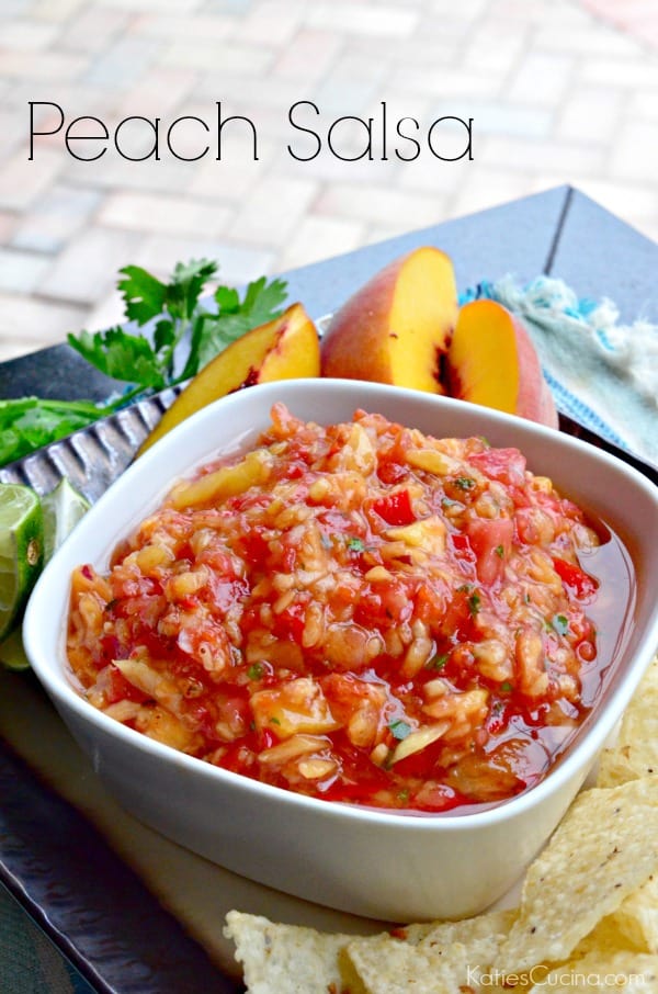 White square bowl holding peach salsa, with fresh peach slices and chips on plate.