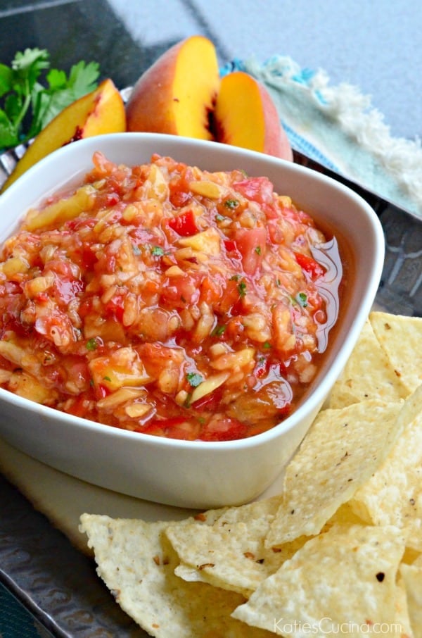 Square white bowl filled with peach salsa on a plate with chips.