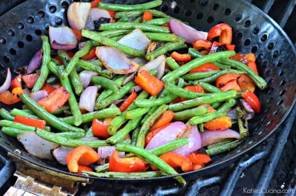 Black grill with a grill basket filled with grilled green beans, red bell peppers, and red onions.