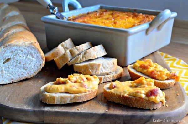 Square baking dish with dip resting on a wood cutting board with sliced baguette and pimento cheese dip. 