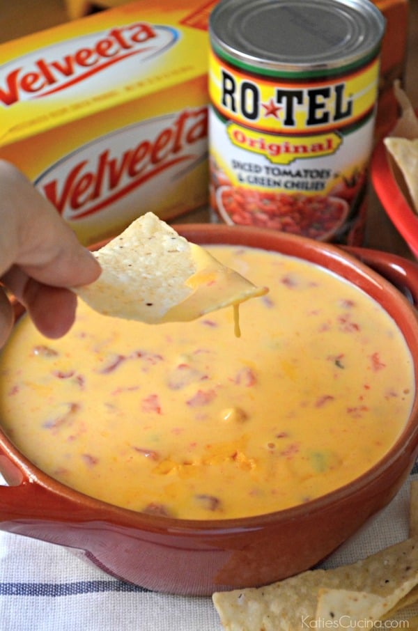 Hand dipping tortilla chip in red bowl filled with queso. 