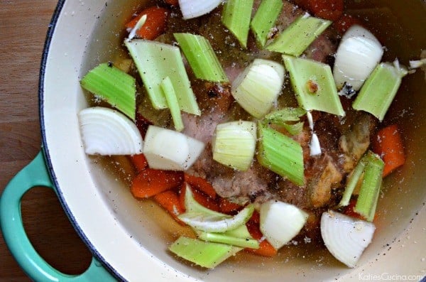White pot with green handle filled with beef bones, chopped carrots, onions, and celery in water.