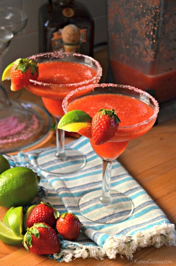 2 Frozen Strawberry Margaritas with sugar and a blender and sugar in the background. 