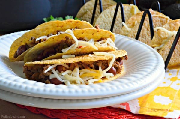 Three crunchy tacos with beef and cheese stacked on two white plates.