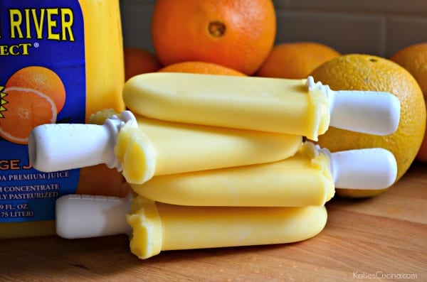 Four stacked orange popsicles with orange juice and oranges in the background.