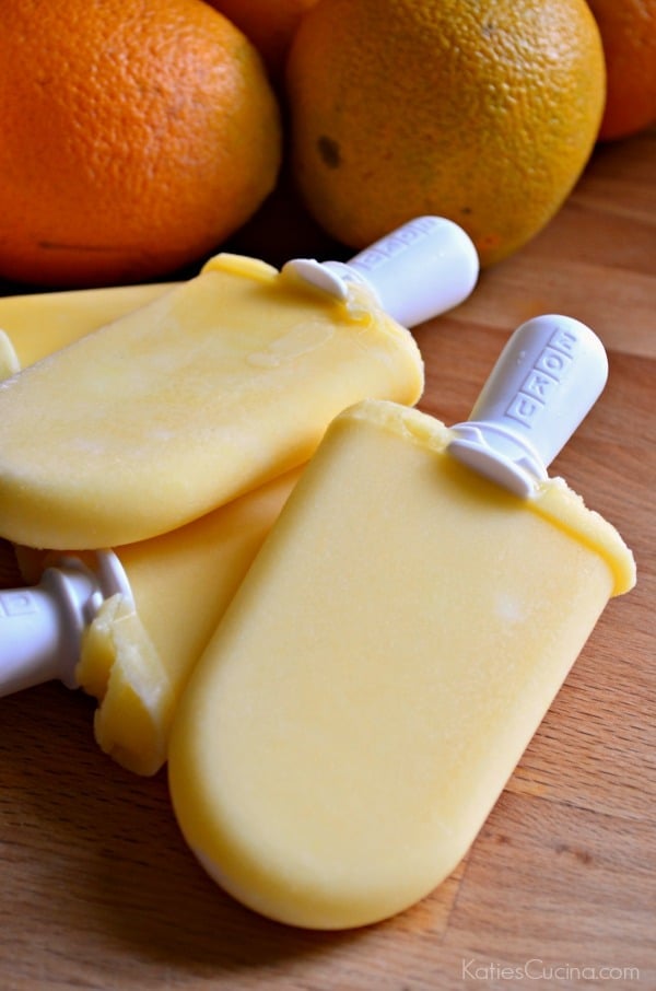 Four orange popsicles bundled together on a wooden countertop with oranges in the background.