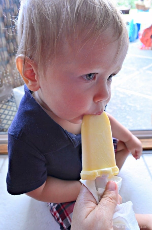 Baby boy sitting down eating an orange popsicles that an adult hand is holding.