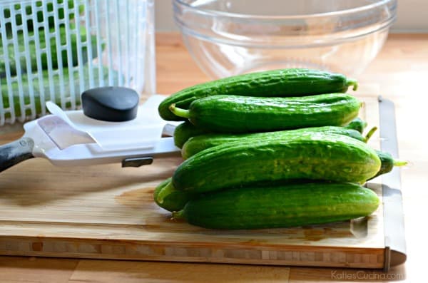 A stack of mini cucumbers on a wood cutting board with a mandoline and produce bin in the background.