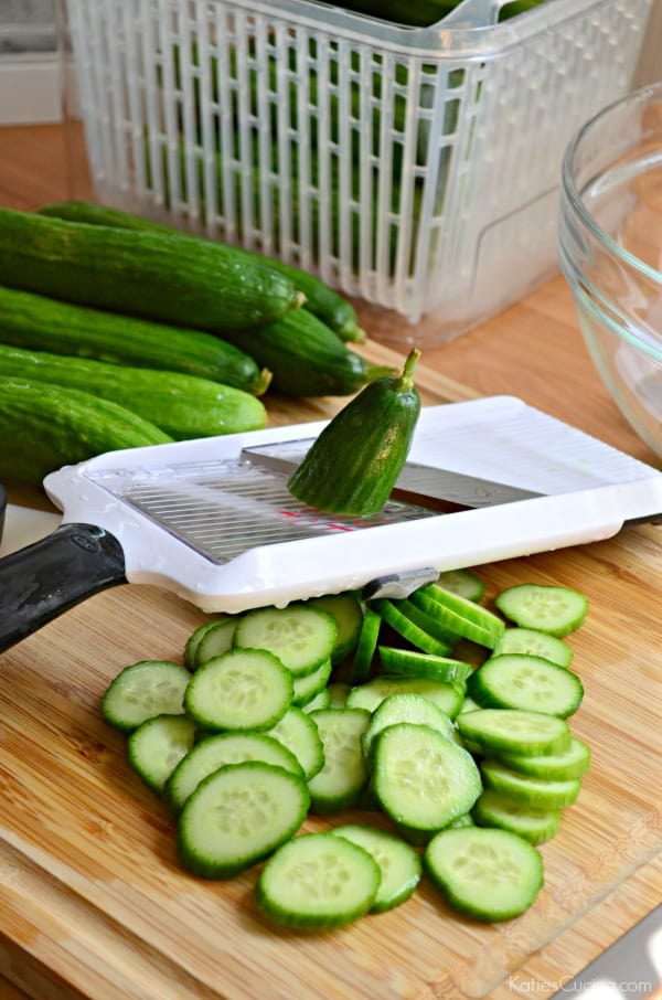 Handheld mandoline slicer with trimmed mini pickles on a wood cutting board.