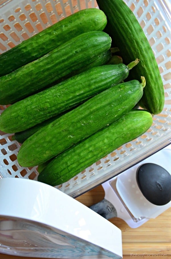 Top view of a white basket filled with whole mini cucumbers on a wooden cutting board.