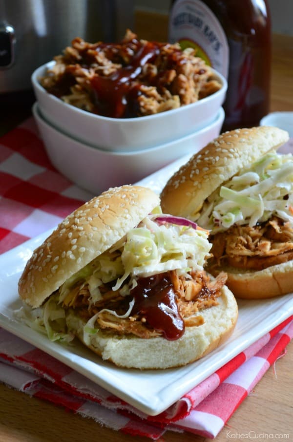 Two pulled chicken sandwiches on a white platter topped with barbecue sauce and coleslaw. 