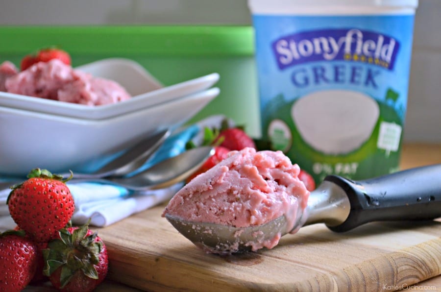 Ice cream scoop with frozen strawberry yogurt on the scoop resting on a wood cutting board. 