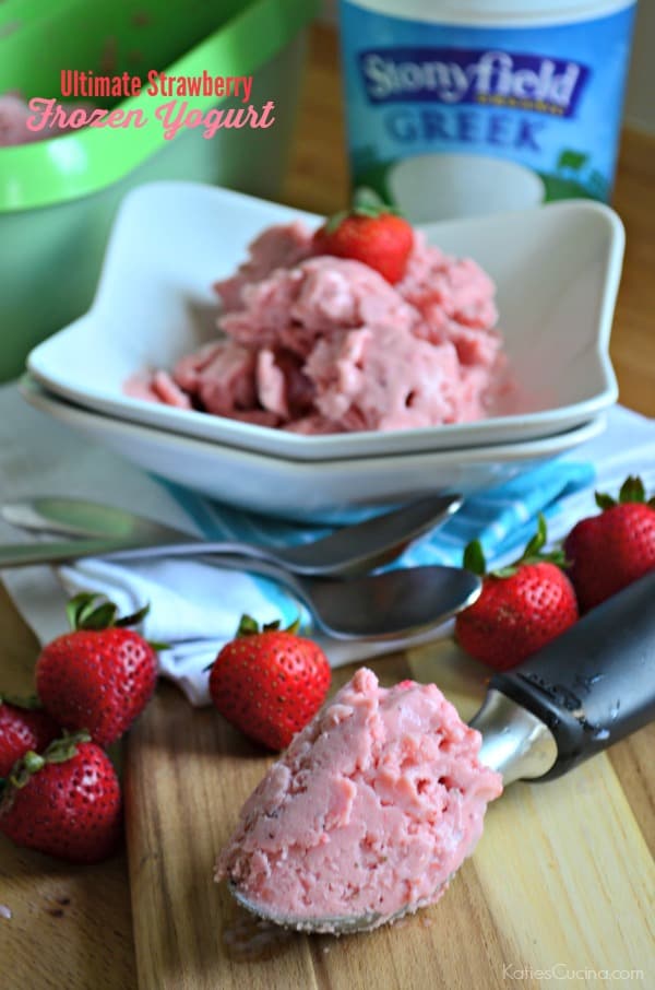 Ice cream scoop filled with Strawberry Frozen Yogurt on a wood cutting board with fresh berries and a bowl of ice cream in background.