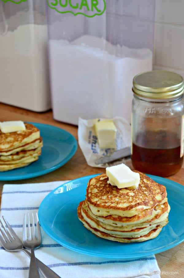 Two blue plates with pancakes stacked on top with butter, syrup, and sugar in the background.