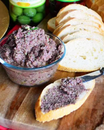 Bowl of brown olive spread with baguettes and spread on one slice.