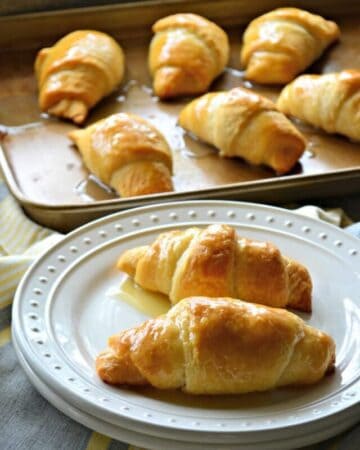 two honey butter crescent rolls on a white plate with a tray of six crescent rolls in the background.