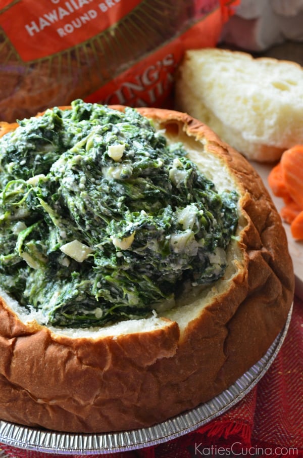 Close up of a bread bowl filled with spinach dip with bread and carrots on the side. 