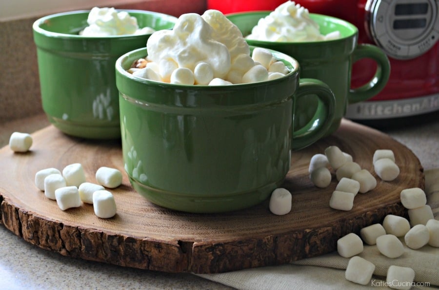 Three green mugs sitting on a slice of wood filled with whipped cream and marshmallows on the counter.