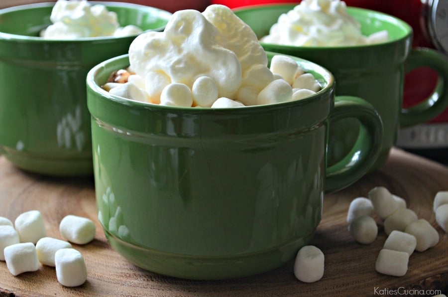 Close up of three large green mugs on a wood slice with mini marshmallows on top.