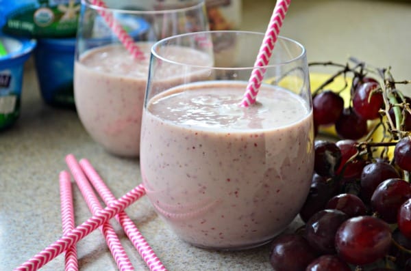 Two clear glasses with light pink smoothie, fresh grapes, bananas, and paper straws.