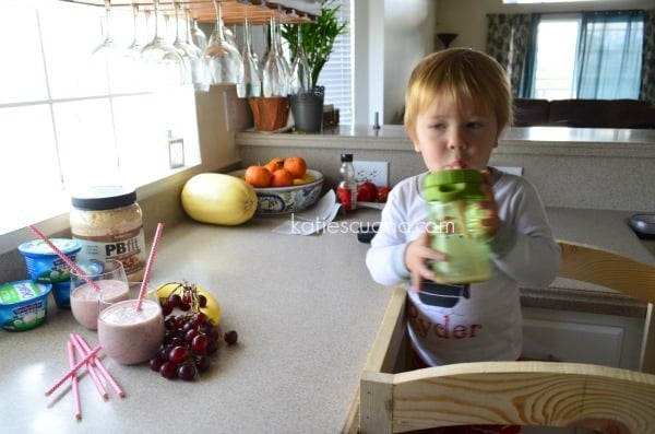 Baby standing on a wood platform by kitchen counter drinking a smoothie.
