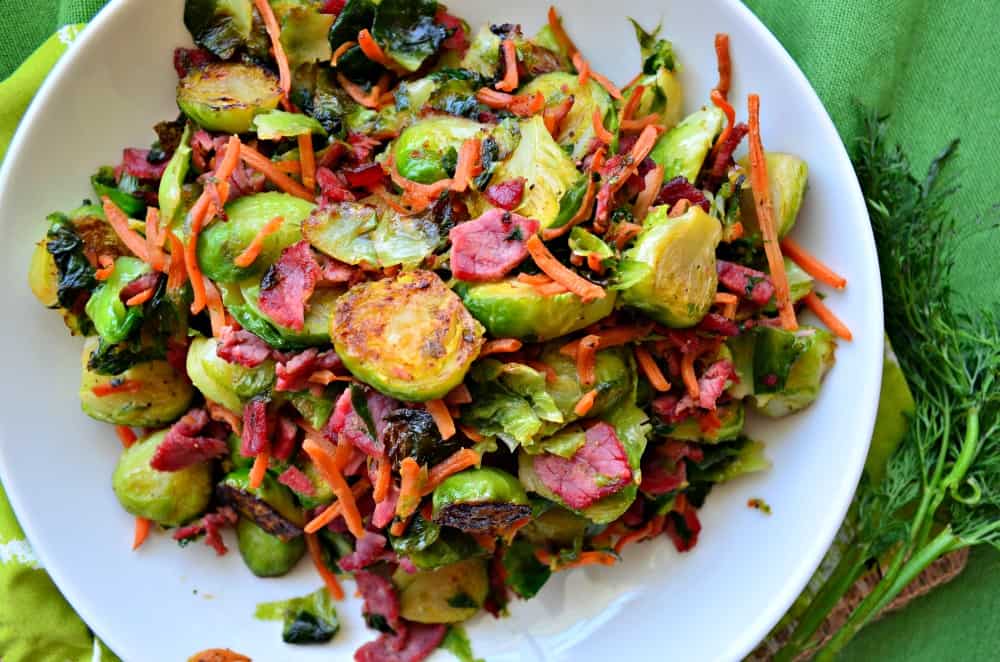 Top view of a white bowl with brussels sprouts, shredded carrots, bacon, and corned beef.