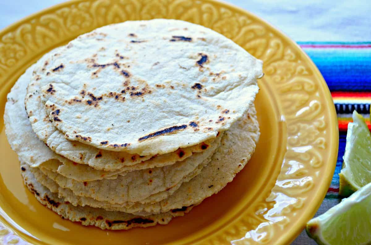 Stack of homemade tortillas on a yellow plate sitting on a colorful cloth.